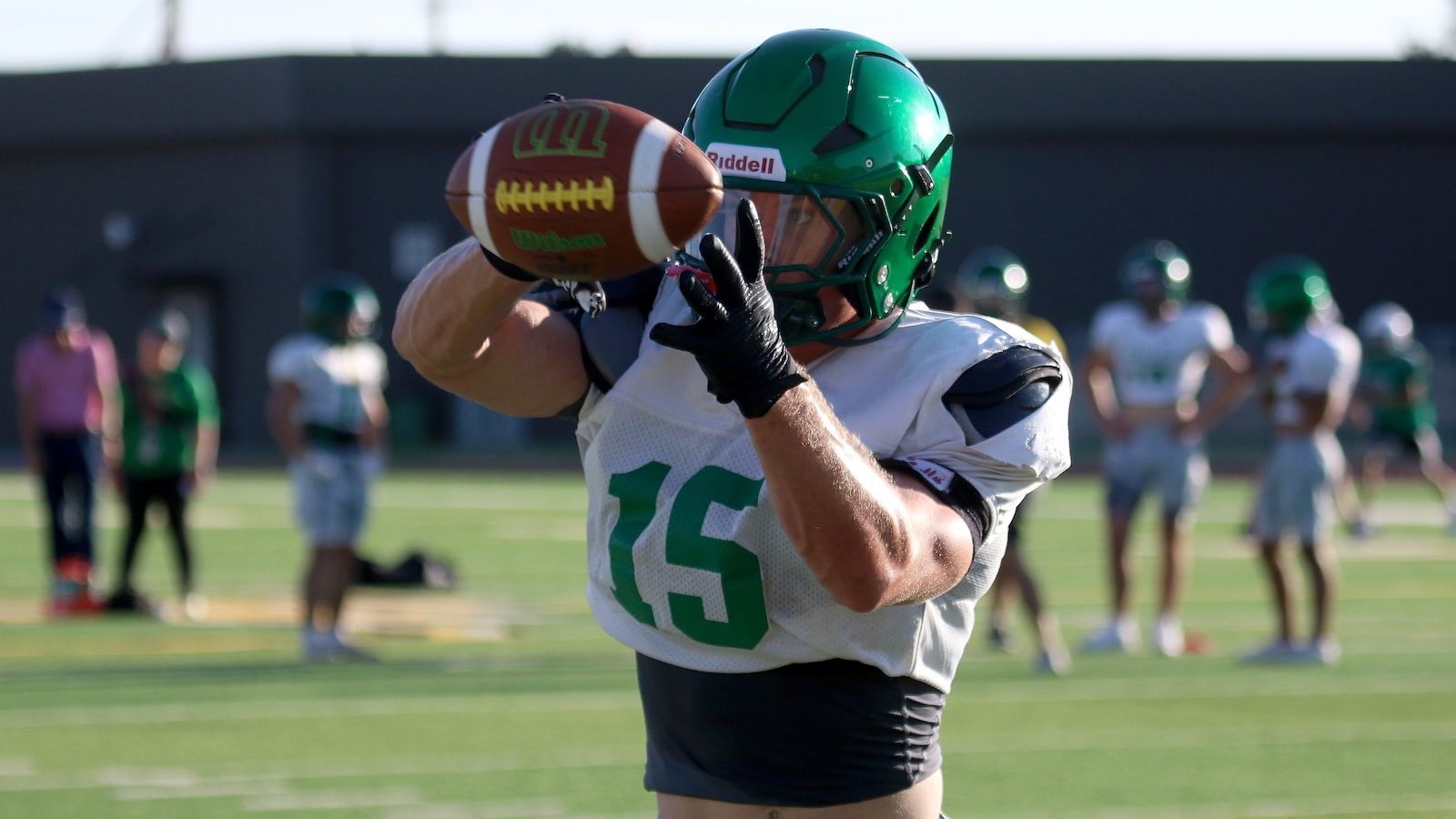 American football player in a white jersey with green number 15 catching a football mid-air on a practice field, green helmet on.