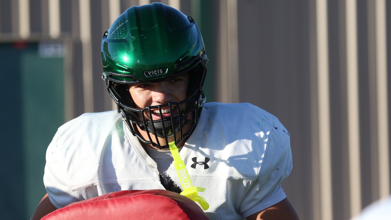 American football player wearing a green Vicis helmet and white jersey, holding a red tackling pad during practice.