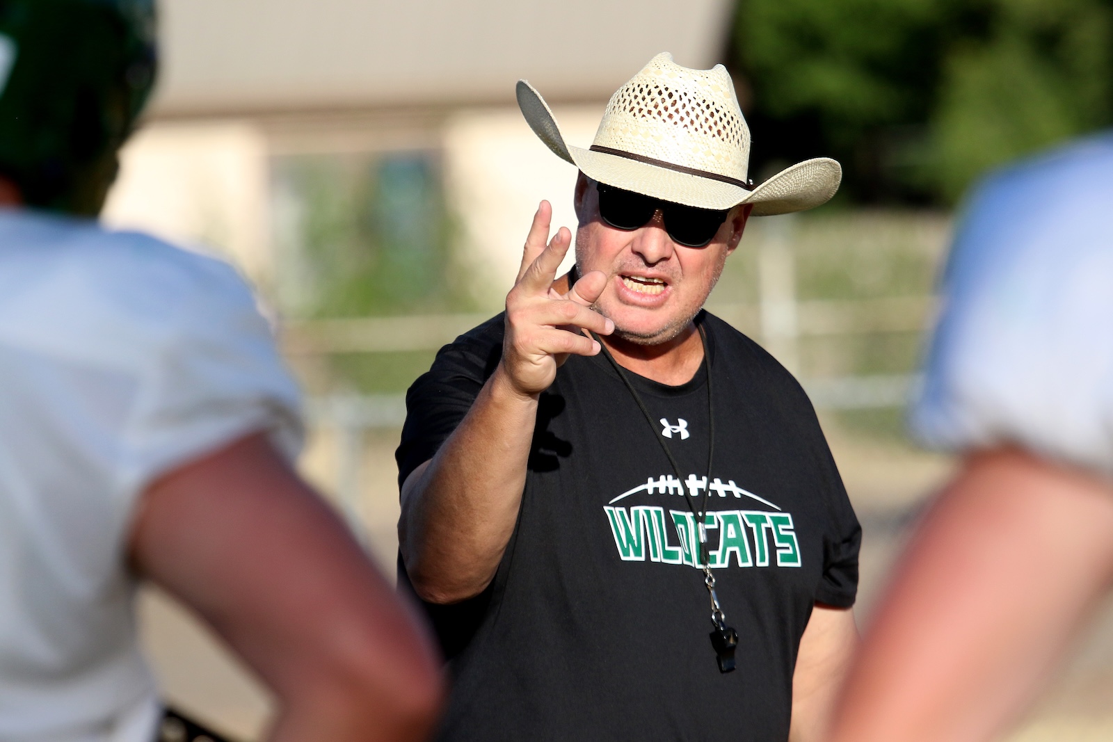 Coach wearing a straw hat and sunglasses points at the camera during a football practice; his shirt says WILDCATS.