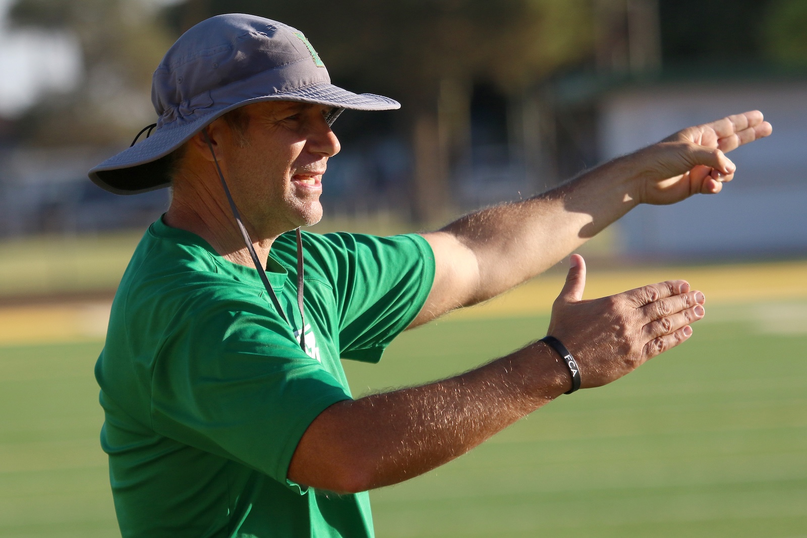Man in a green shirt wearing a wide-brim hat, signaling with his arms extended on a grassy field.