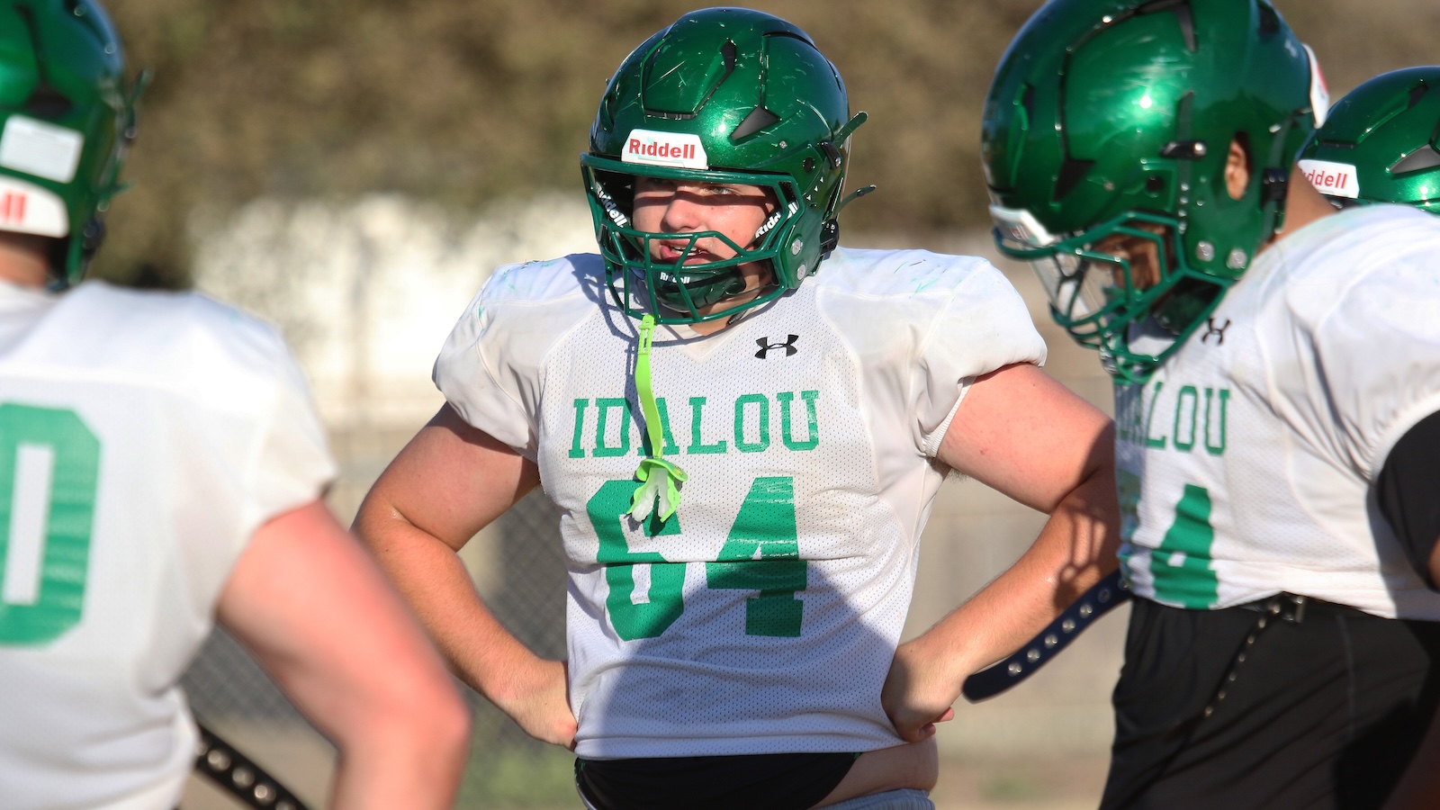 Football players in green helmets and white IDs jerseys with'IDALOU' on the chest, huddling on a practice field.