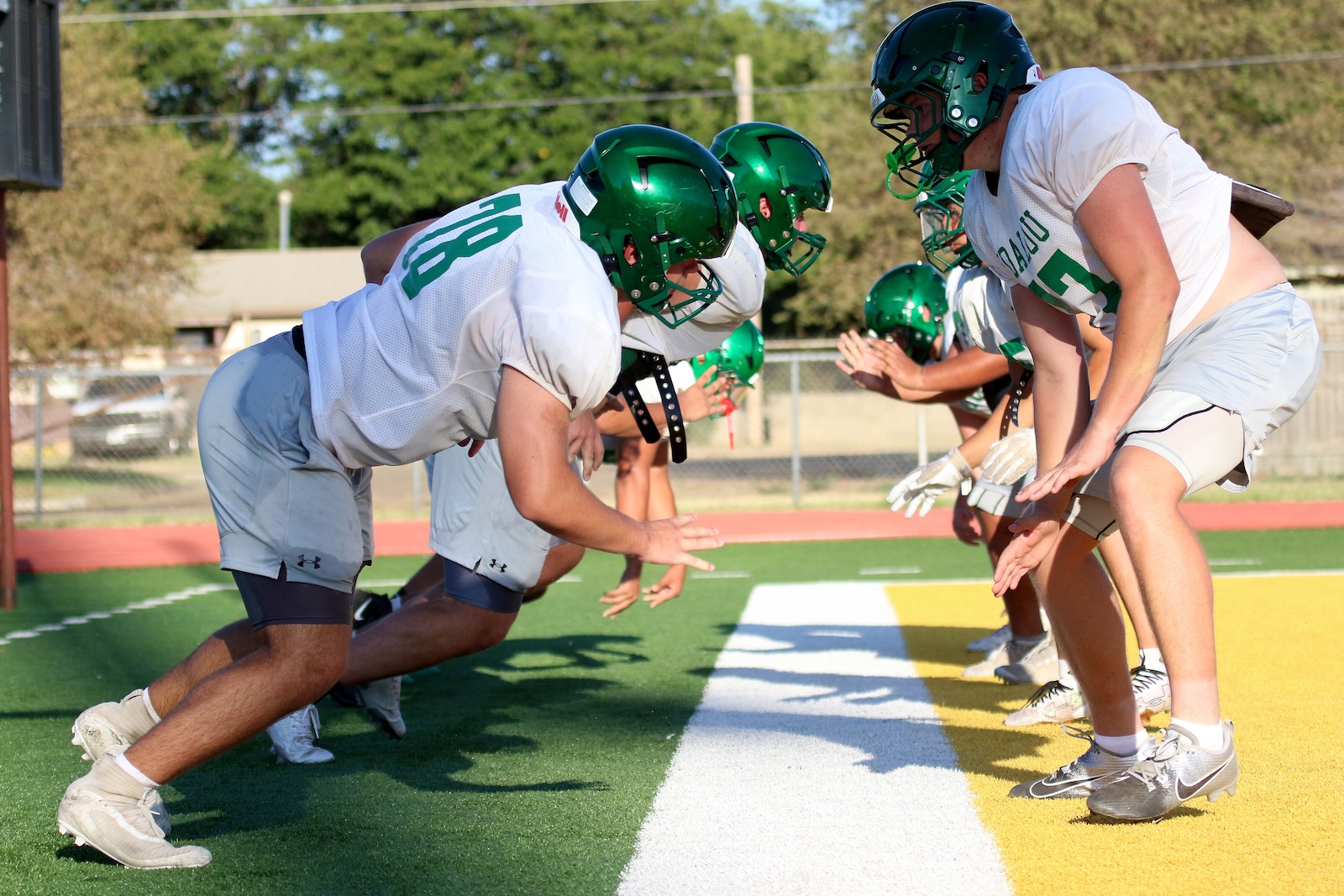 Football players in white uniforms line up in a scrimmage stance on a green artificial turf field with green helmets facing each other.