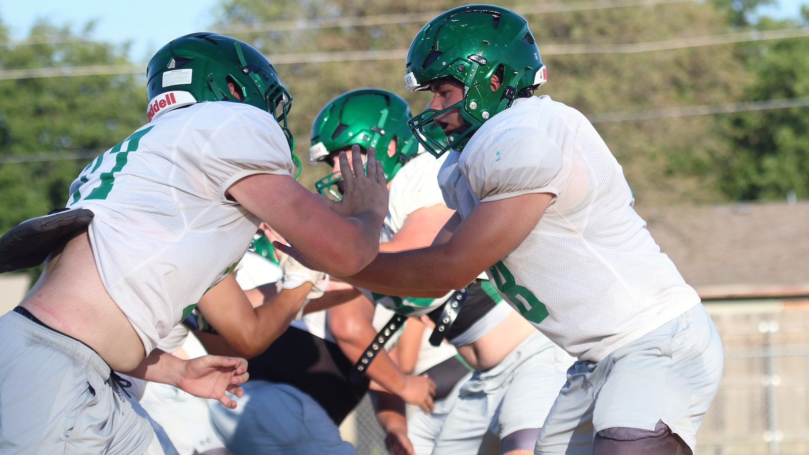 American football players in white uniforms and green helmets engage in a blocking drill on a sunny field.