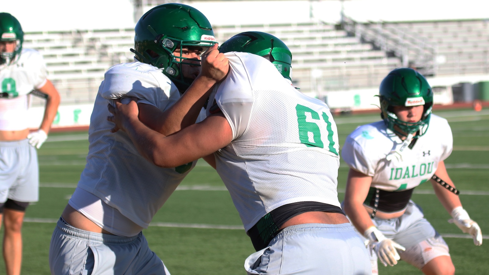 Two American football players in white jerseys and green helmets grapple in a drill on a sunny practice field, number 61 visible on one player's back,