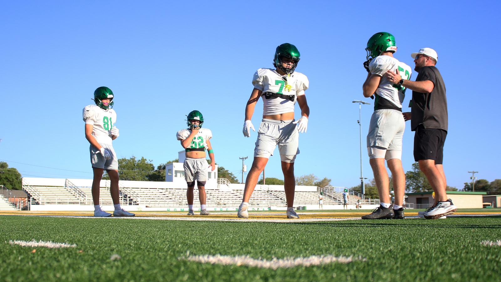 Youth football players in white uniforms and green helmets stand on a sunny practice field as a coach talks to one player. Bleachers and goalposts visible in the background.