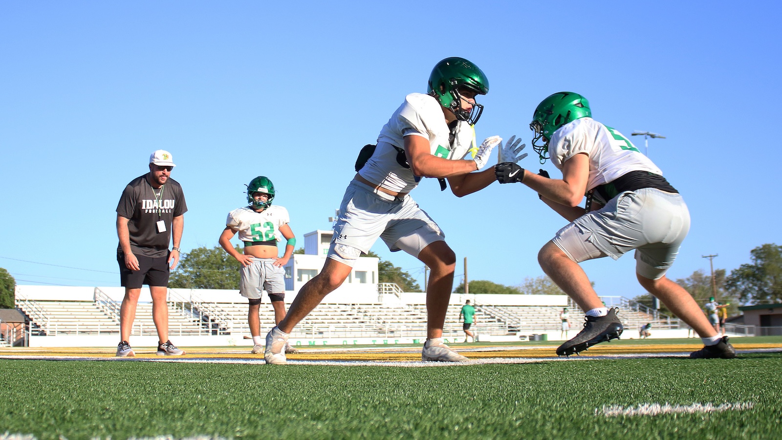 Two football players in white uniforms with green helmets engage in a blocking drill on a sunny field, coach watching in the background.
