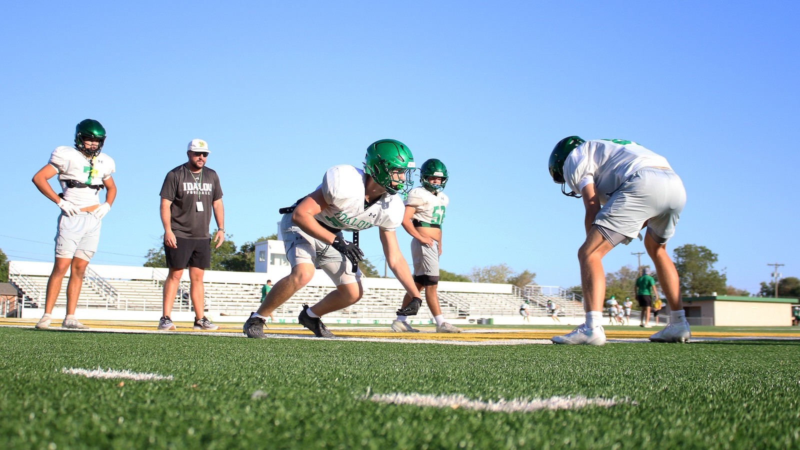 Football practice on a sunny field with players in green helmets lined up and a coach watching.