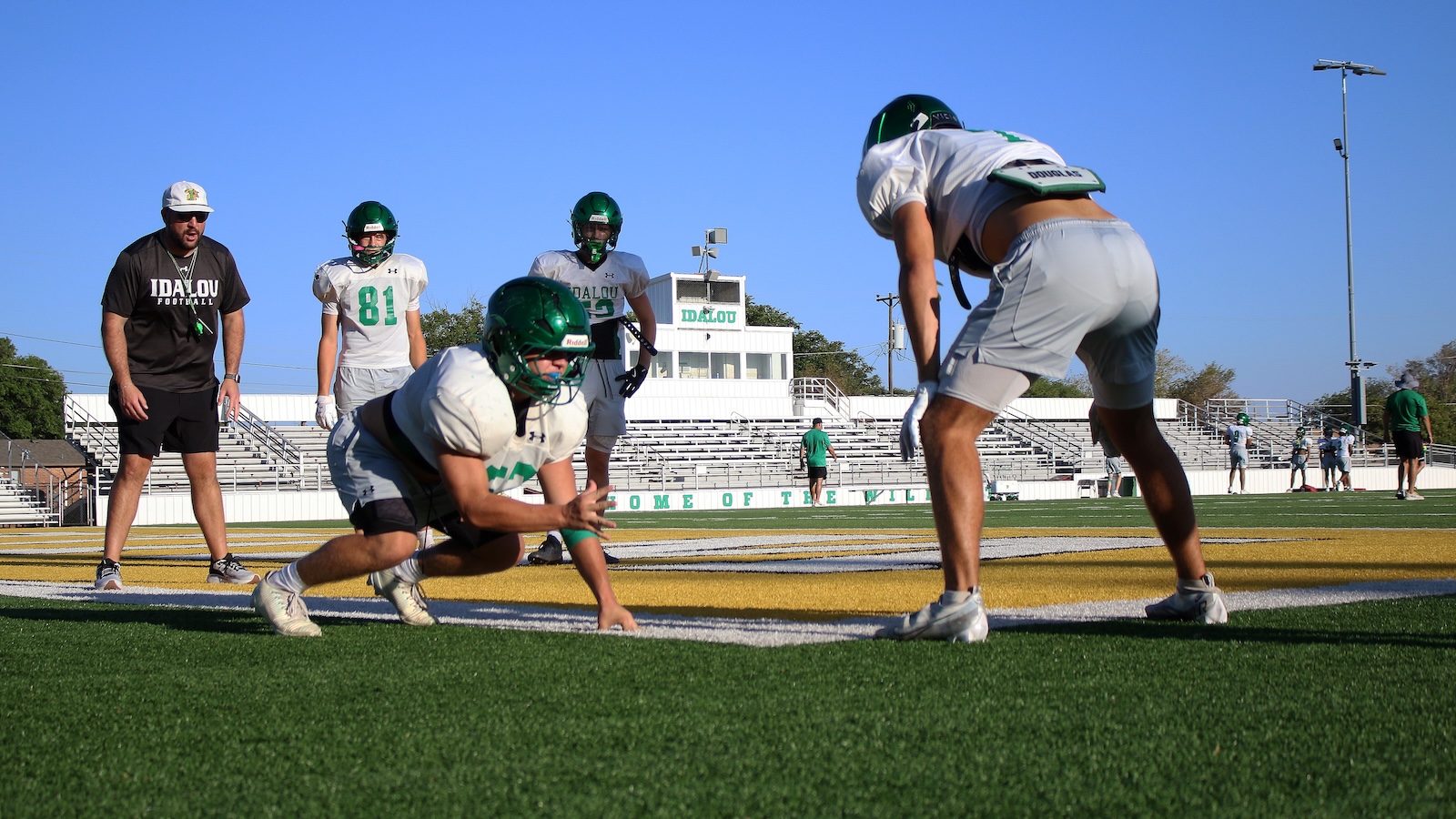 Football players in green and white uniforms on a sunny practice field; one player crouches low on the turf while another stands nearby and watches.