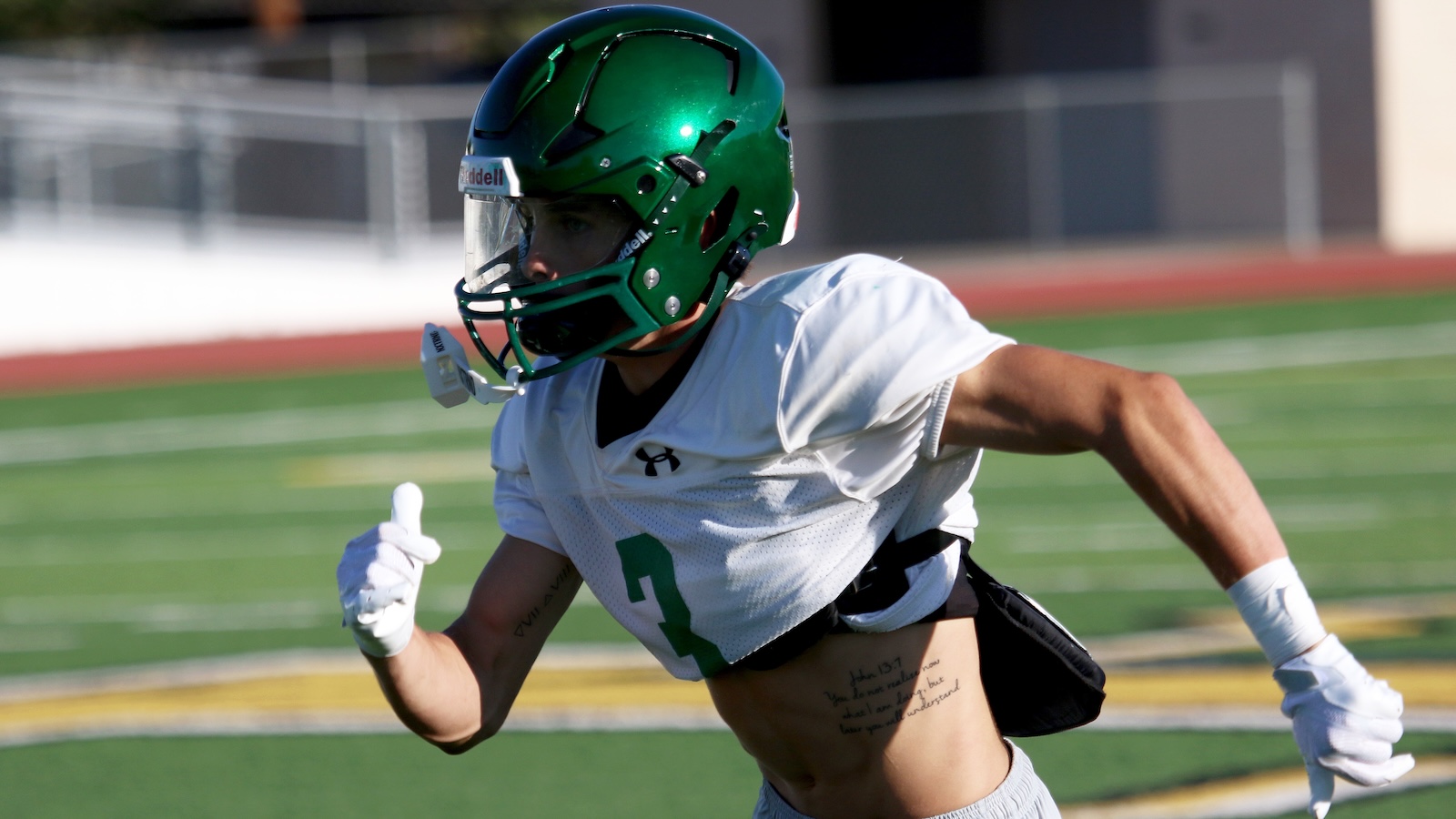 American football player in a white jersey with the number 3 and a shiny green helmet sprinting on a turf field during practice or game.