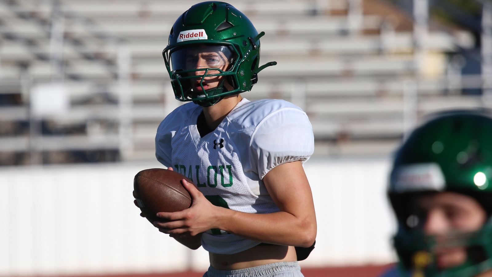 Young football player in a white jersey and green helmet holds a football on the field.