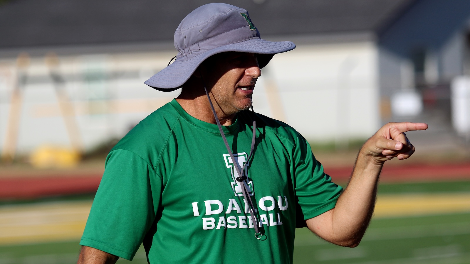 Baseball coach in a green Idaho Baseball shirt points while wearing a wide lavender sun hat on a sunny field.