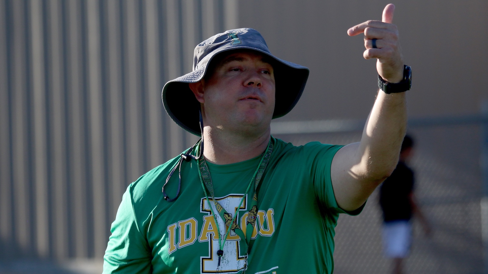 Man in a green Idaho T-shirt and wide-brimmed hat raises his right thumb in a thumbs-up gesture outdoors, with a watch on his left wrist.