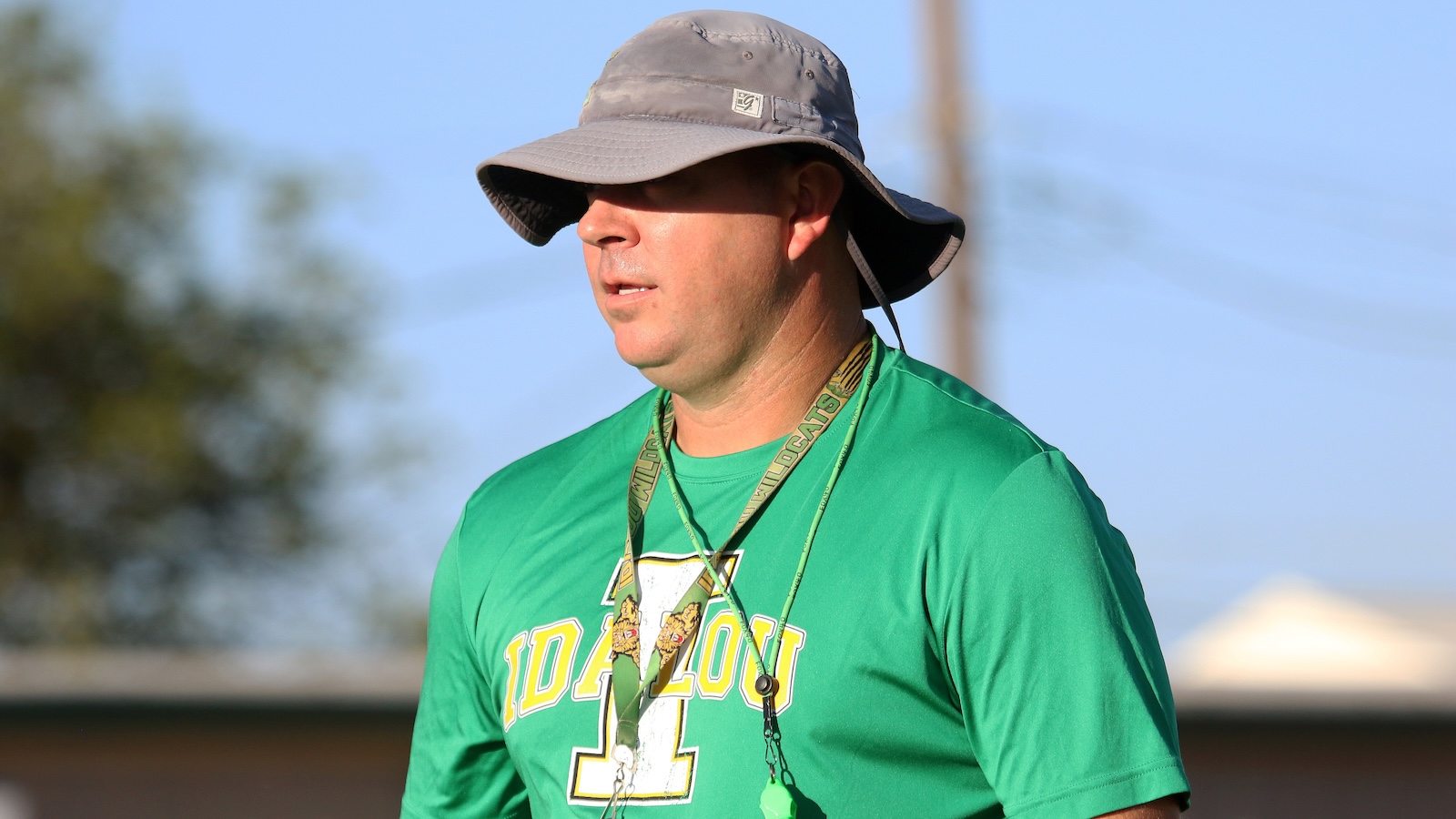 Man in a green Idaho shirt wearing a wide-brimmed gray hat, standing outdoors with a lanyard around his neck.