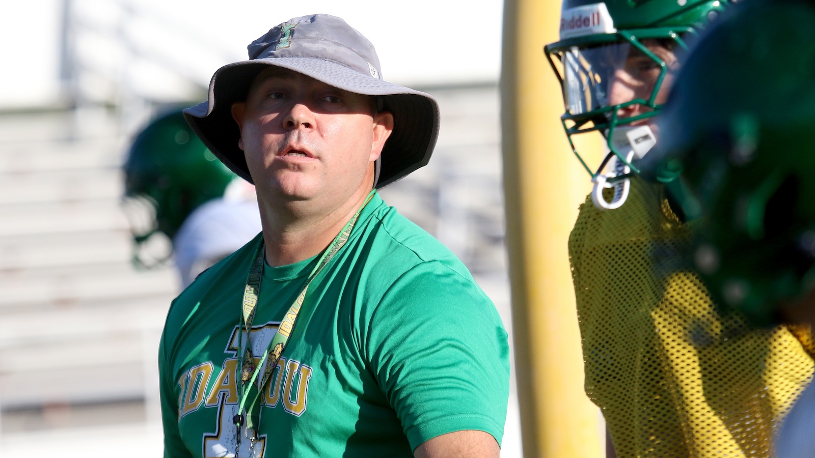 Football coach in a green shirt and gray bucket hat watches practice on the sunny sideline, with players in green helmets nearby.