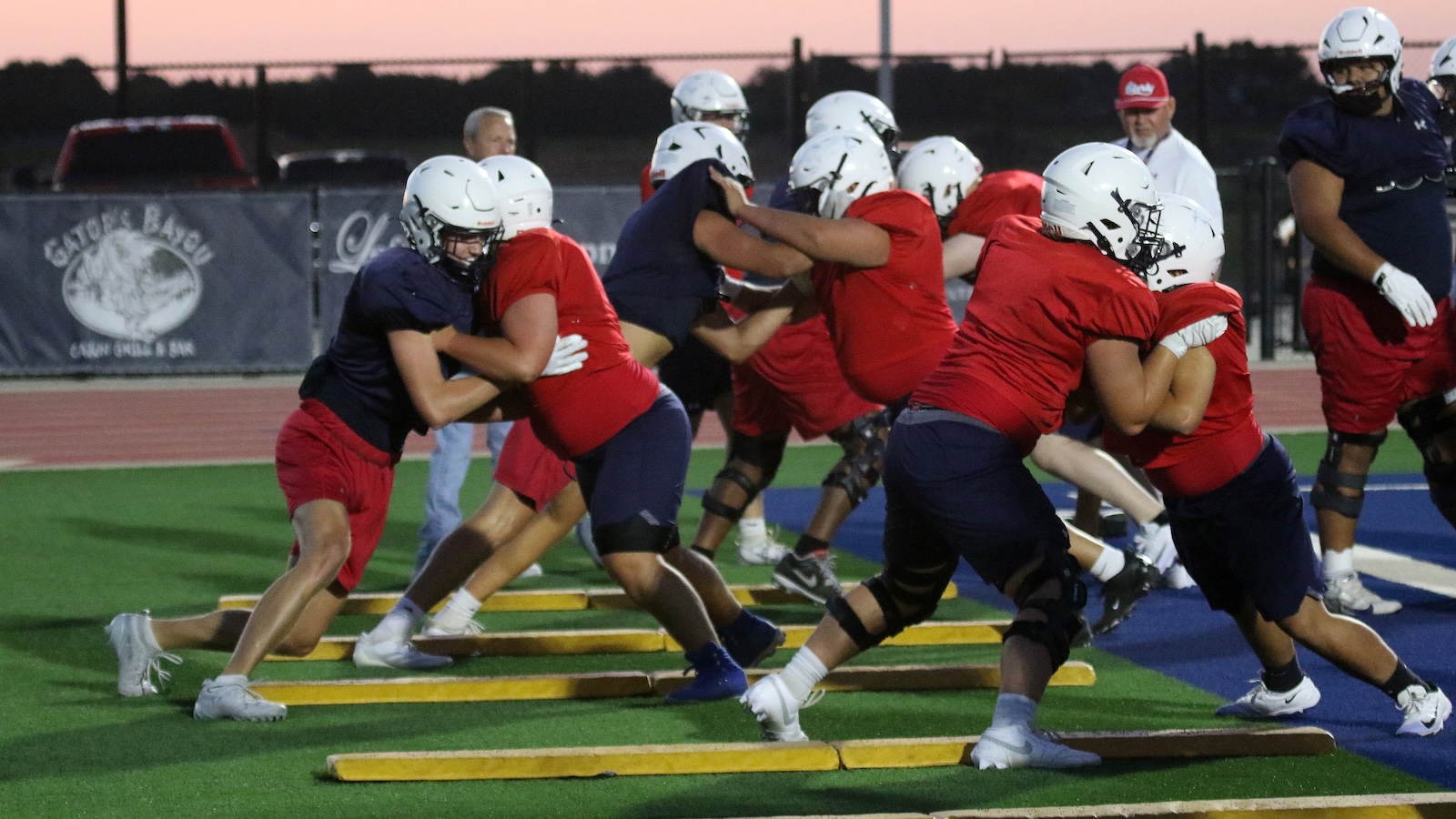 Young football players in red and navy practice jerseys blocking on artificial turf with wooden blocking shields in a line drill at sunset venue.