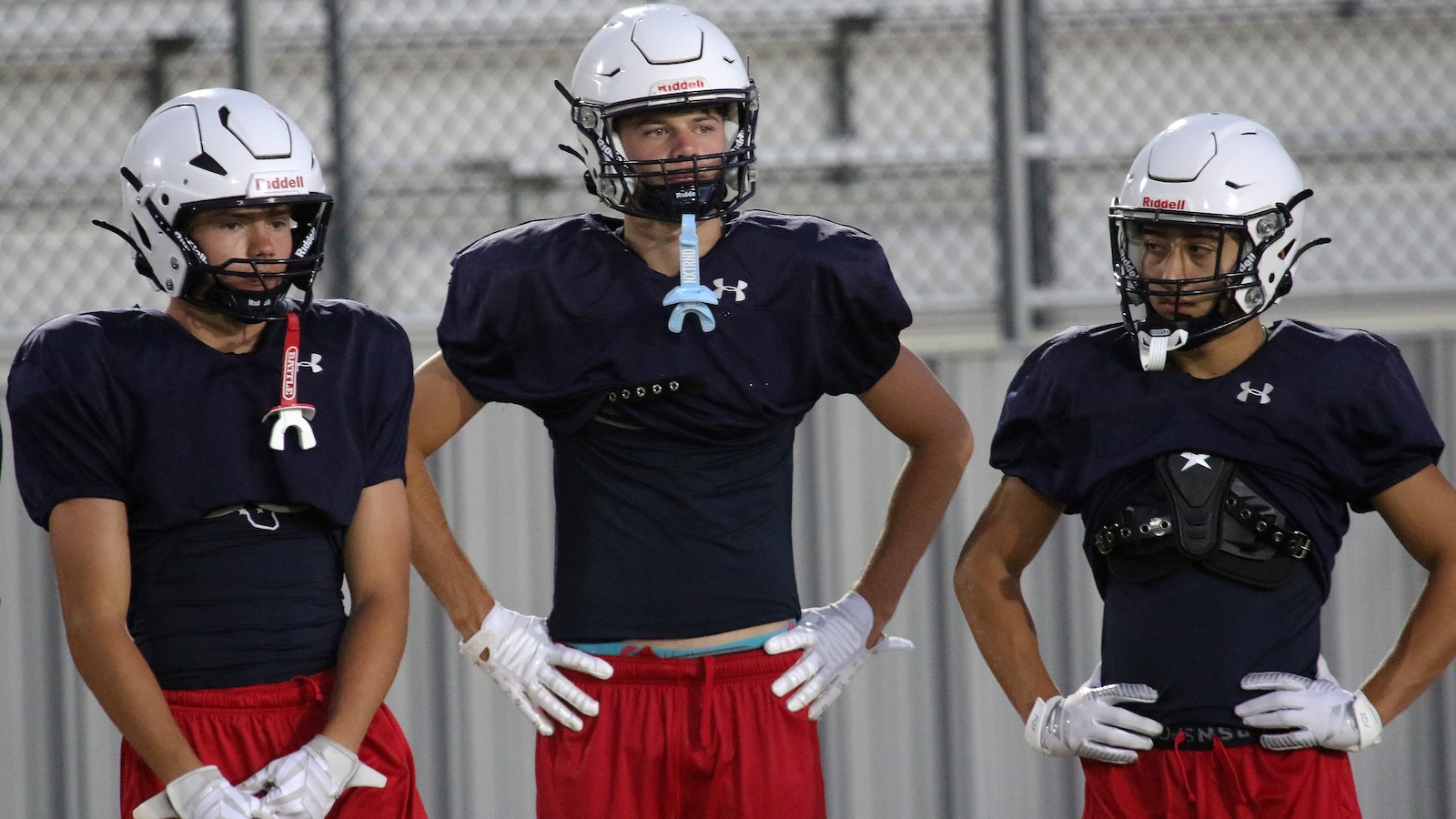 Three football players in navy practice jerseys and red shorts stand with hands on hips during drills on a field with a chain-link fence behind them.