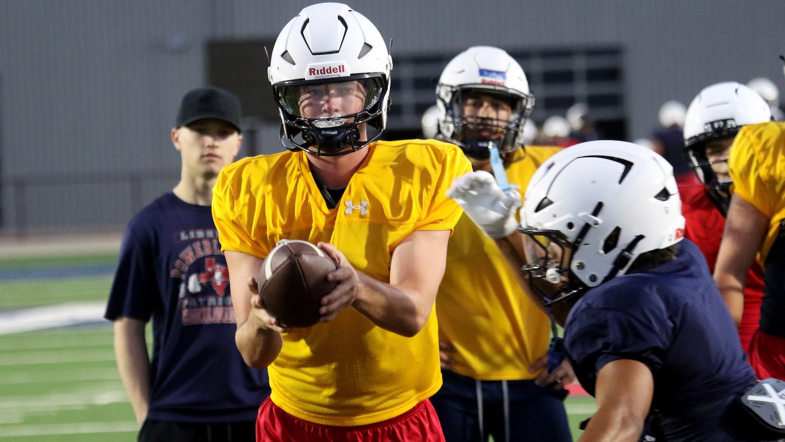 Football practice: a player in a yellow jersey holds a ball, ready to throw, with teammates and a coach in the background.