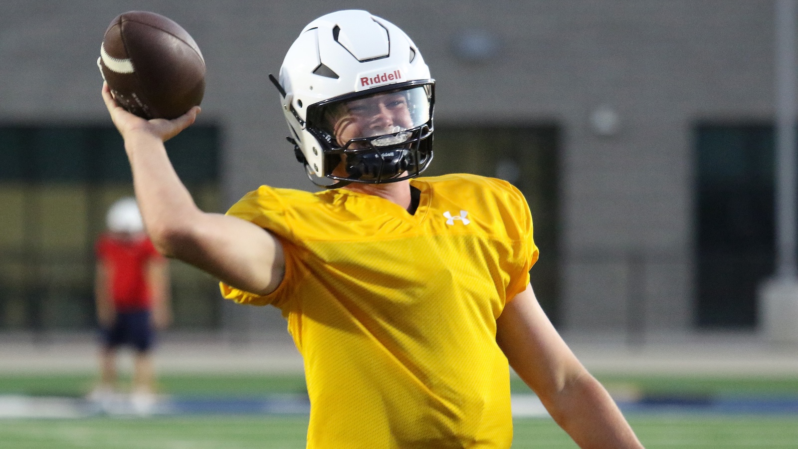 Quarterback in a yellow practice jersey throwing a football, wearing a white Riddell helmet on the field.