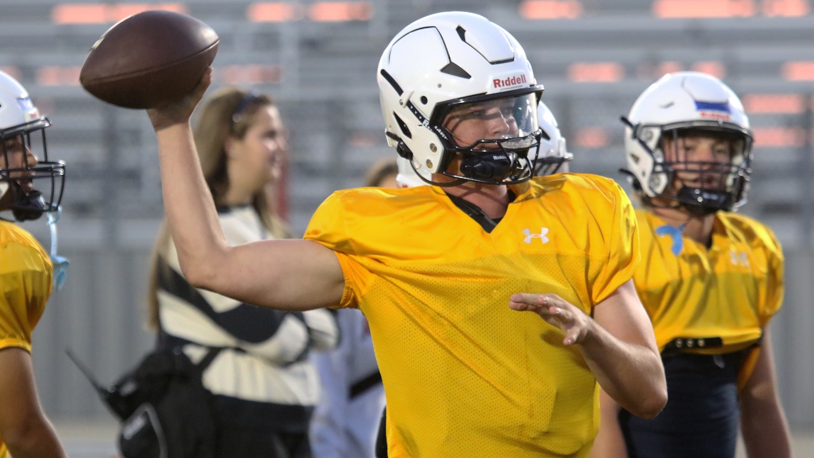 Quarterback in yellow jersey throws a football during practice on a field with teammates in the background.