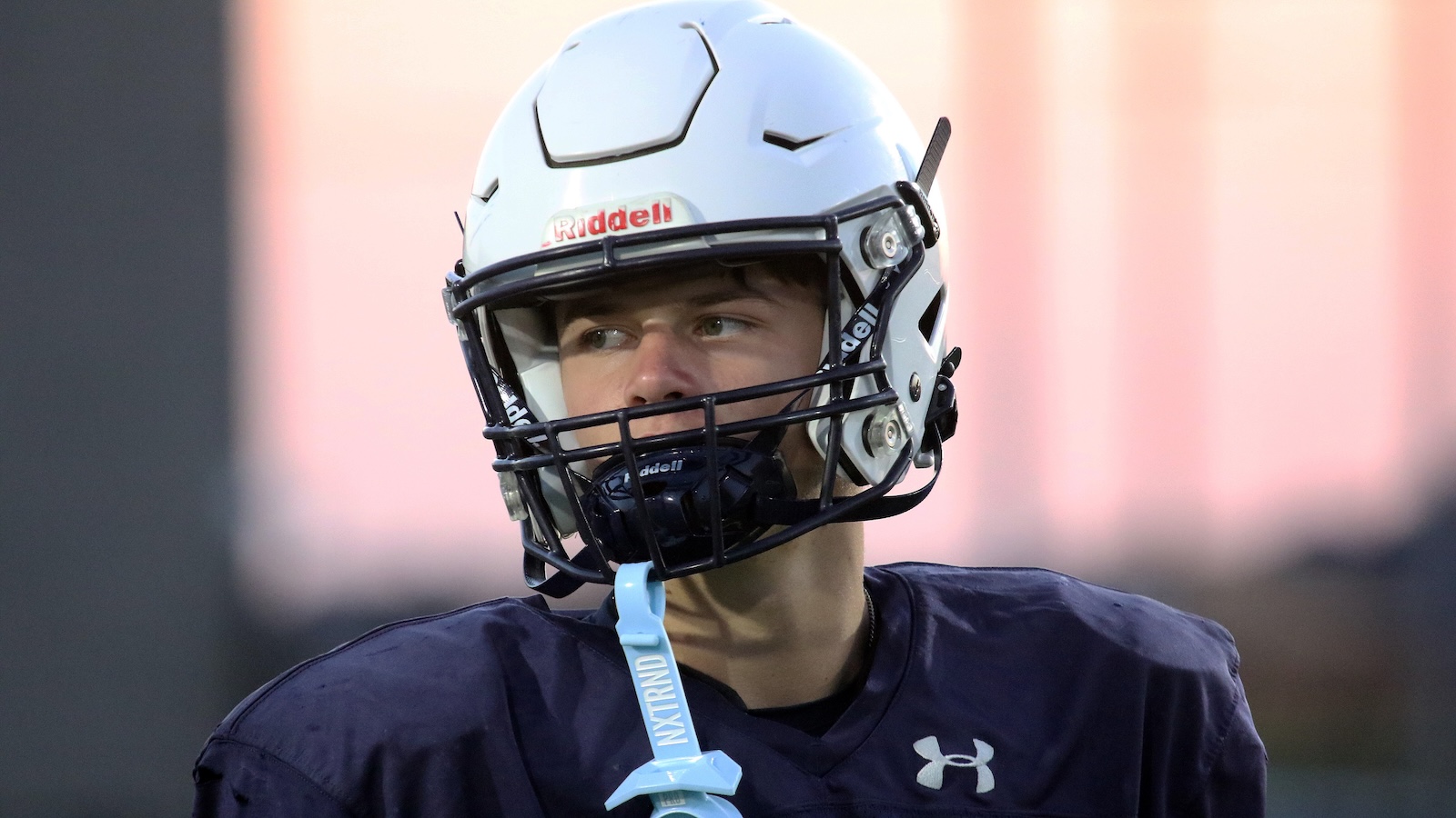 Football player in a white helmet and navy jersey looking to the right, with a blue mouthguard and lanyard visible.