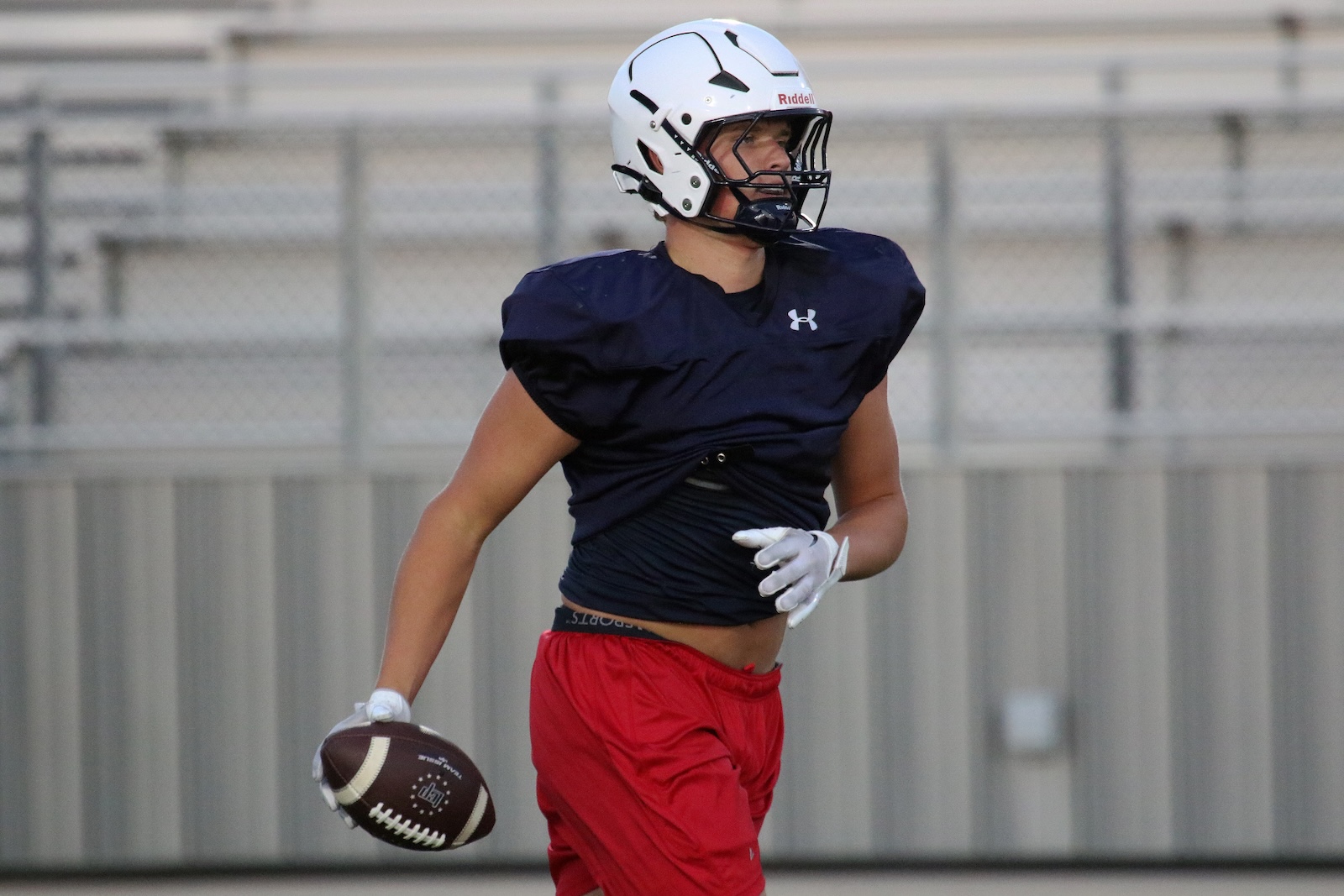 Football player in navy jersey and red shorts runs with a ball during practice, wearing a white helmet and gloves.