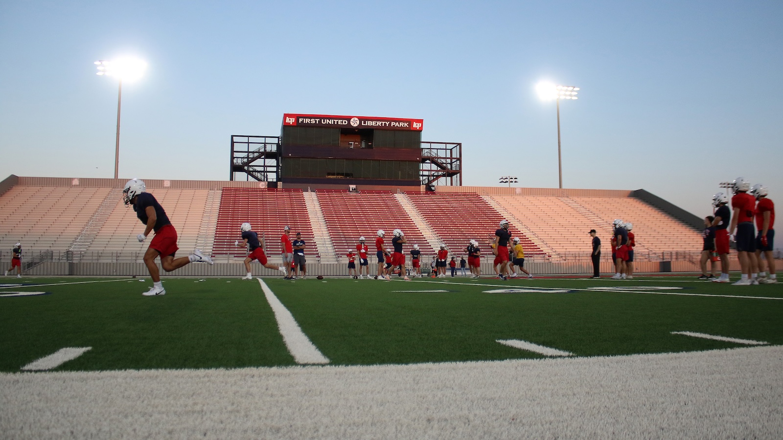 Group of football players in helmets practicing on a green field at dusk, empty red bleachers in the background with bright stadium lights on.
