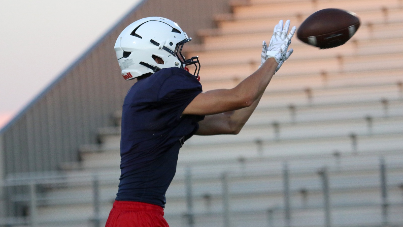 Football player in a navy jersey and red shorts reaches to catch a pass, wearing a white helmet and gloves, with empty stadium bleachers in the background.