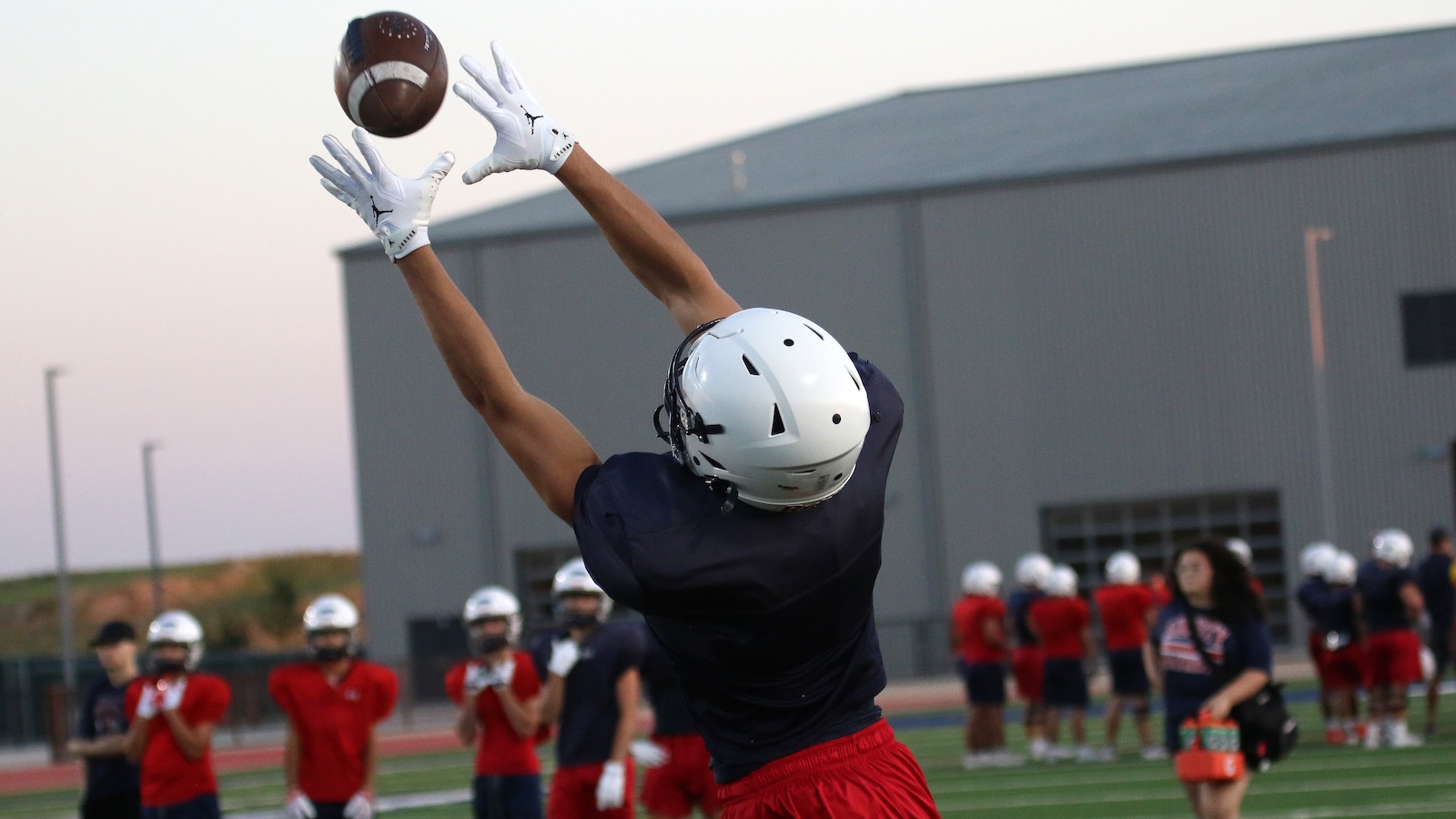 Football player in navy shirt and red shorts reaches to catch a ball mid-air with white gloves during practice on a field.