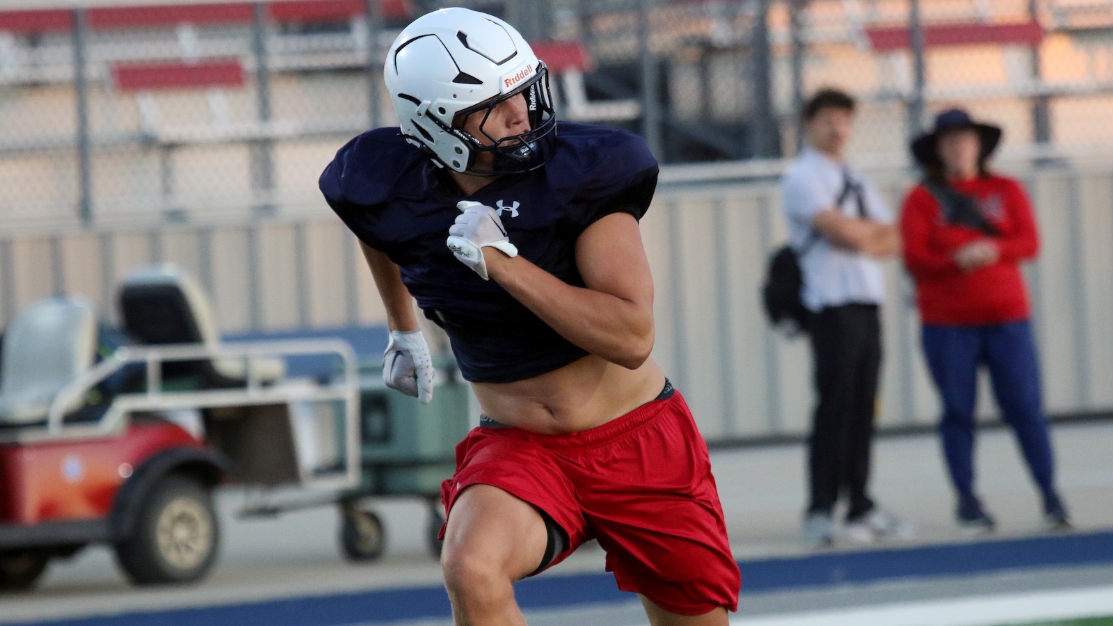 Football player in a navy jersey and red shorts runs on a field, wearing a white helmet and gloves.