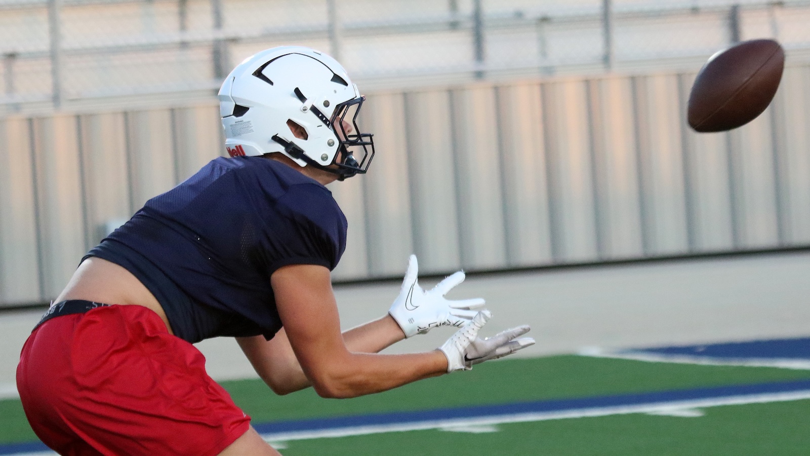 Football player in a navy jersey and red shorts lunges to catch a pass, wearing a white helmet and gloves on a turf field.