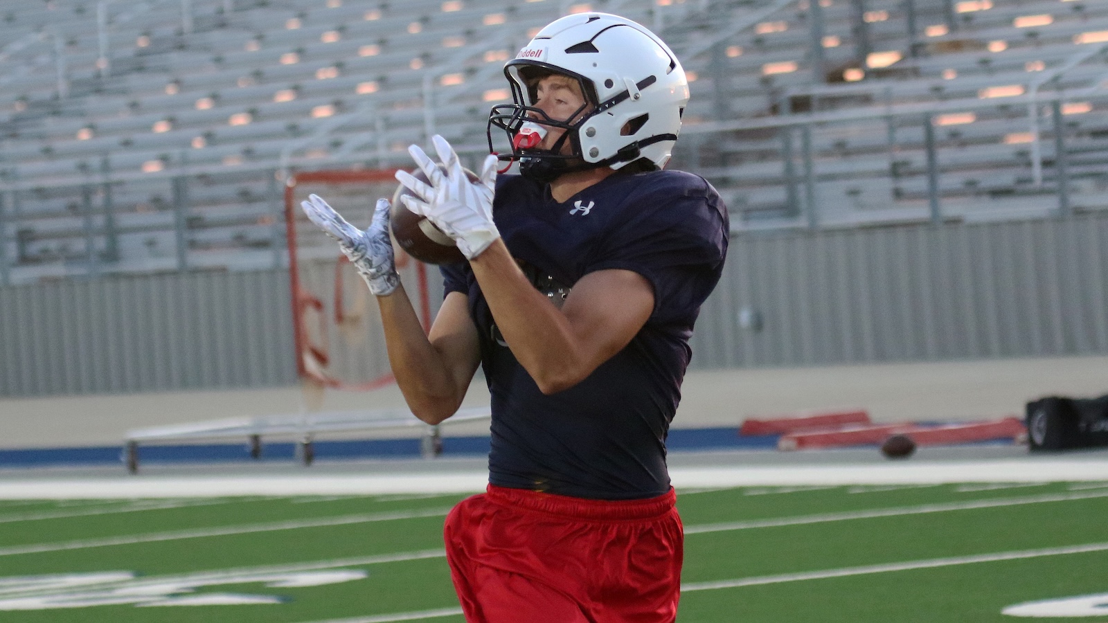Football player in a navy jersey and red shorts catches a pass on a turf field with a white helmet on.