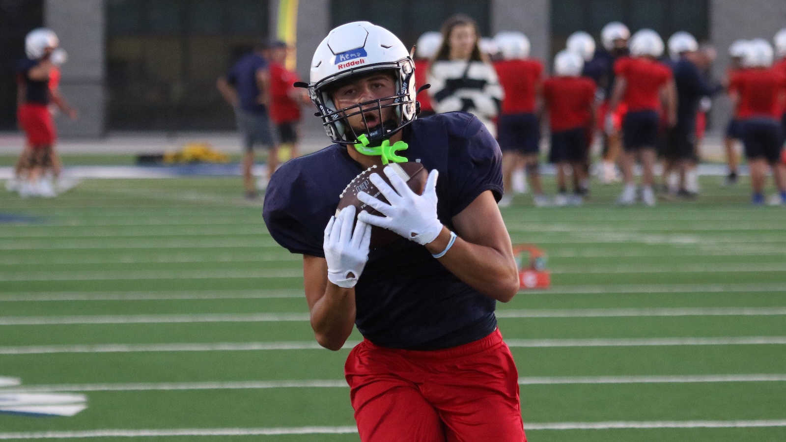 Football player in navy jersey and red shorts runs with a football on a practice field, wearing a white helmet and gloves.