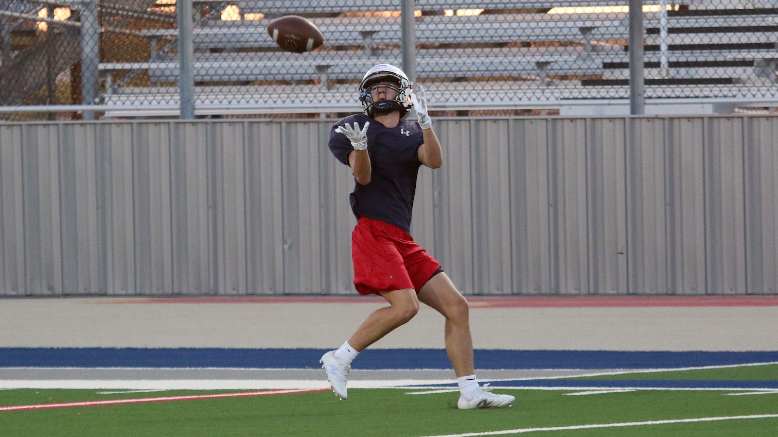 Football player in a navy shirt and red shorts catching a football on a green turf field, wearing a helmet and gloves.