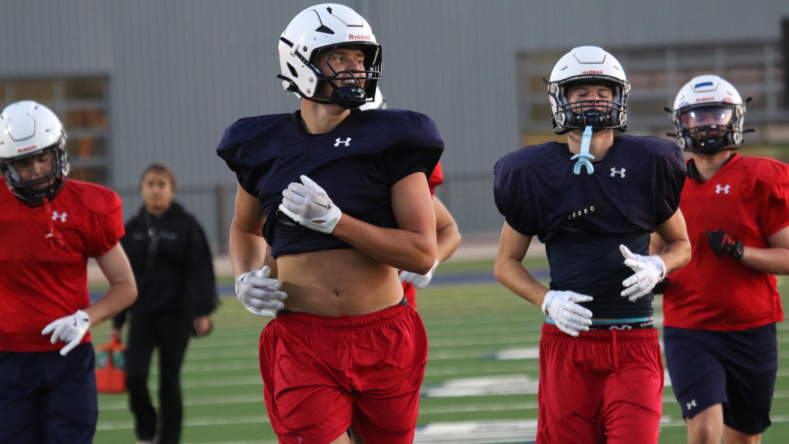 Football players in navy practice jerseys and white helmets sprint on a green field during a drill.
