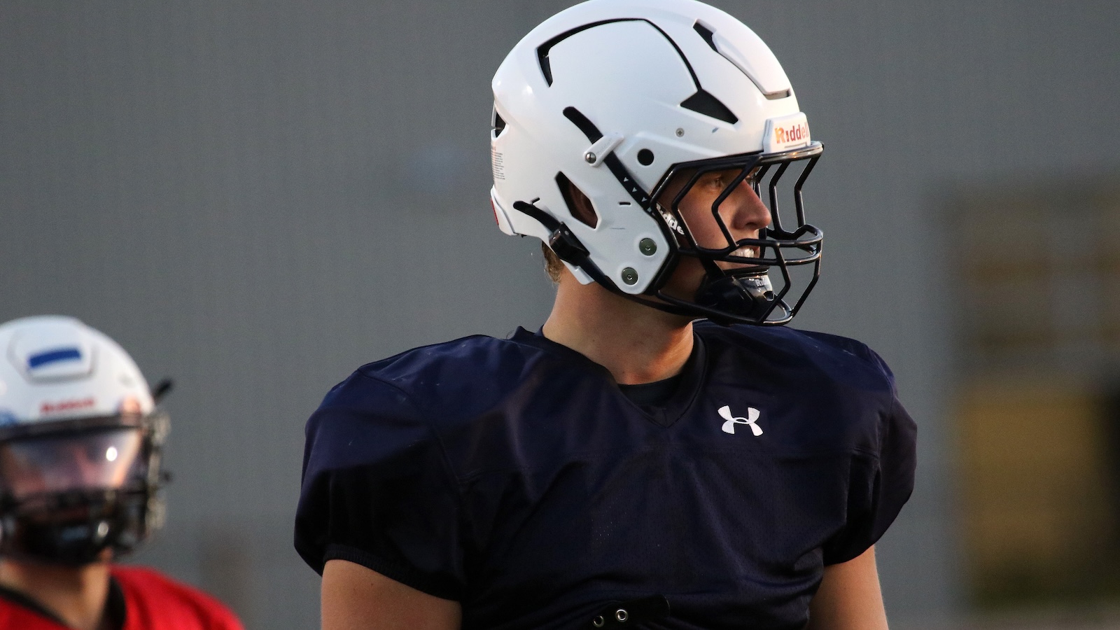 Side view of a football player wearing a white helmet and navy practice jersey, looking to the right on the field.