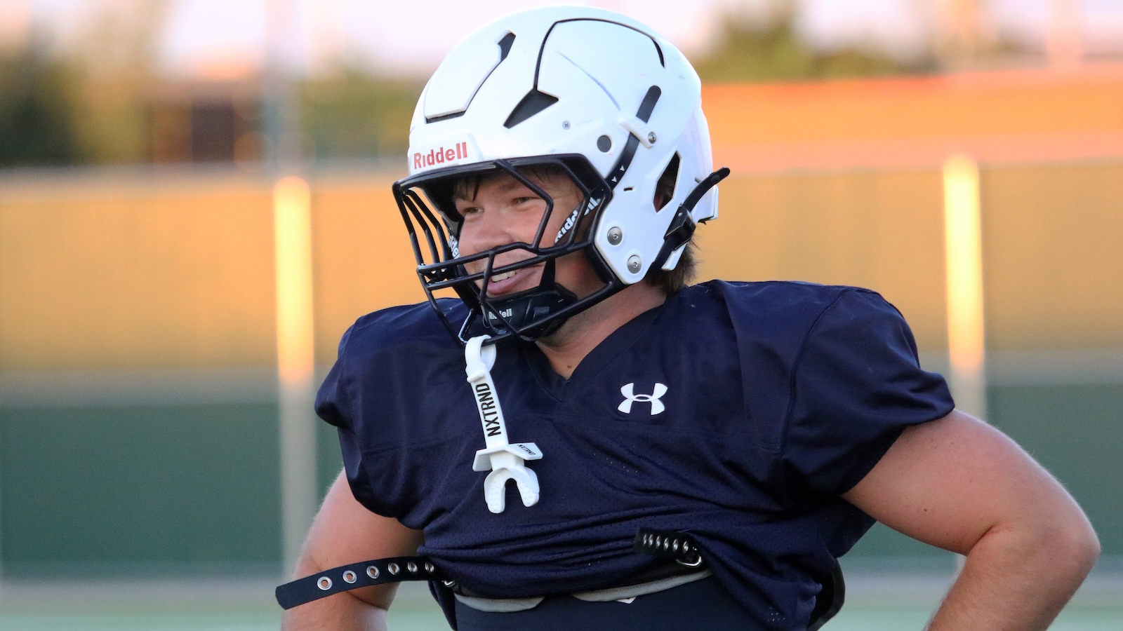 Male football player in a navy practice jersey wearing a white Riddell helmet, smiling on the field with hands on hips.
