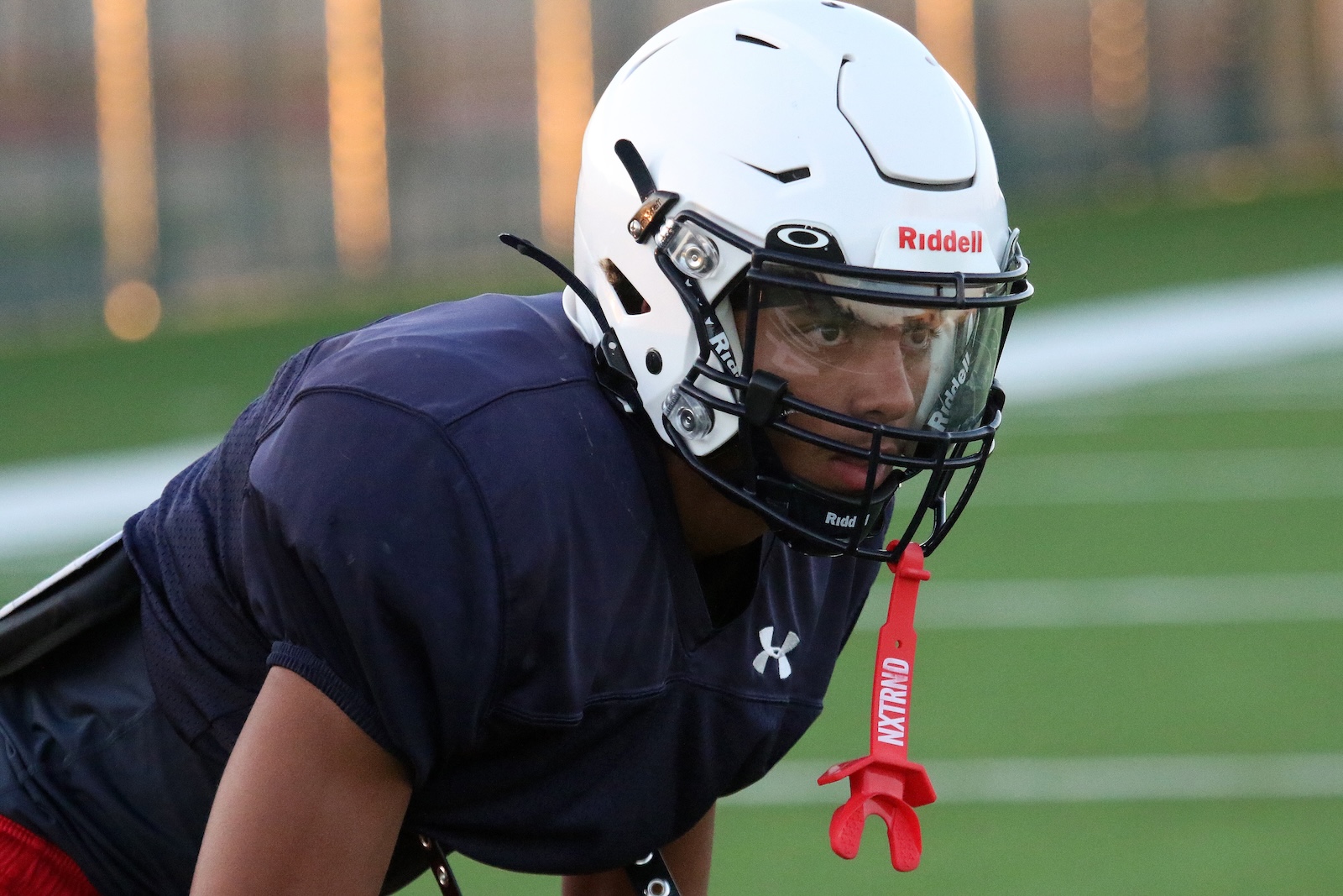 American football player in a white Riddell helmet and navy jersey leans forward on a green practice field during drills, mouthguard tether visible