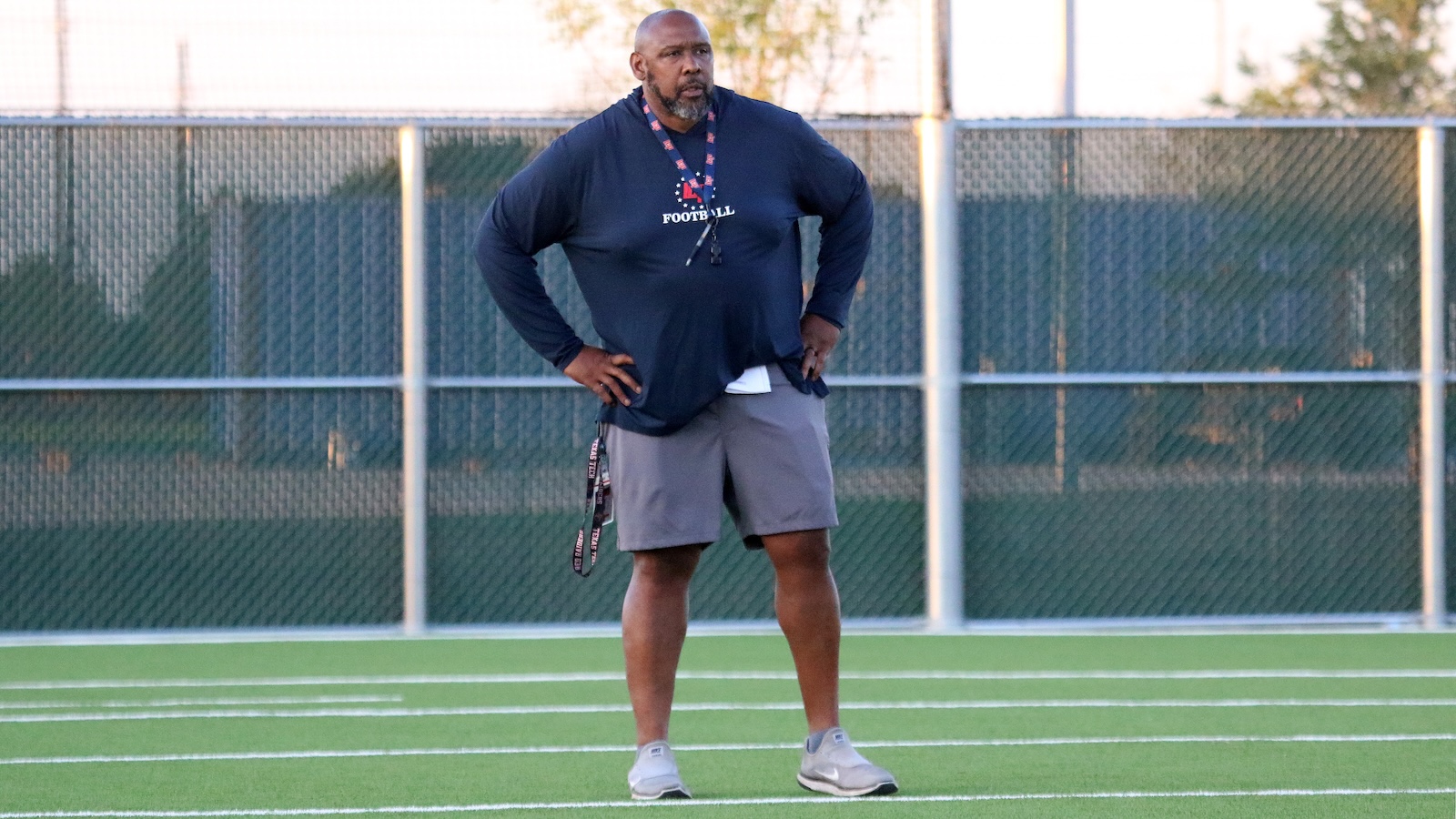 Male football coach standing on a turf field with hands on hips, wearing a navy long-sleeve shirt and whistle lanyard.