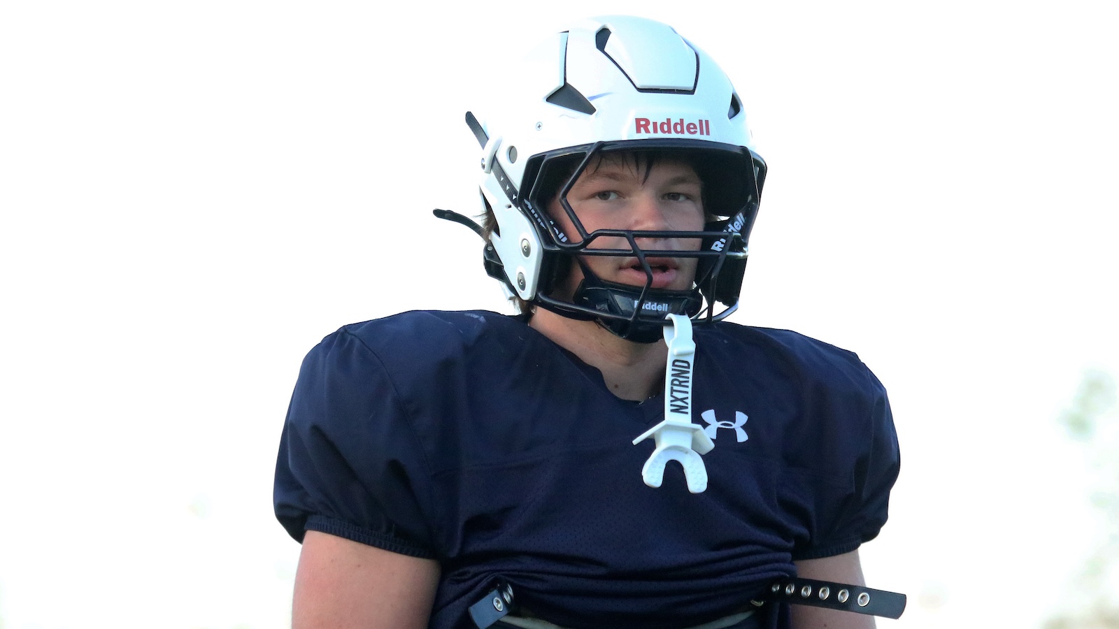 American football player in a navy jersey wearing a white Riddell helmet with a face mask, mouthguard hanging from the mask.