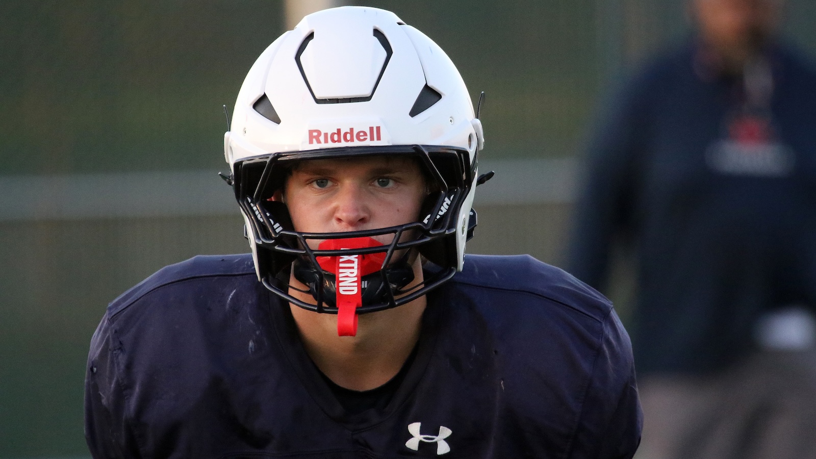 Portrait of a football player in a white Riddell helmet and navy jersey, red mouthguard visible at practice field