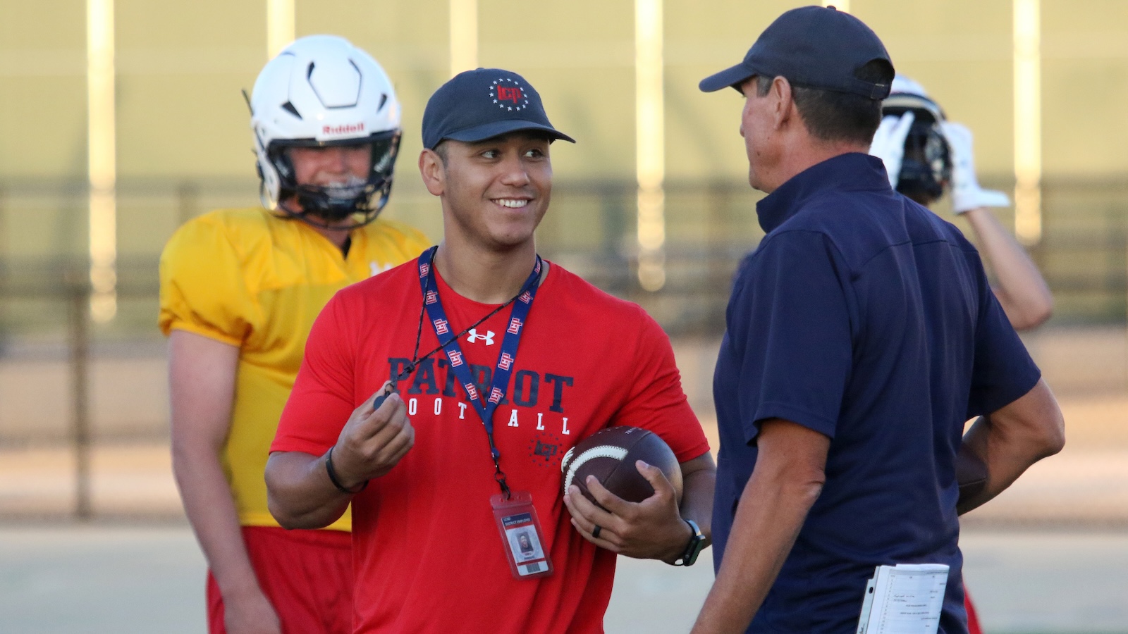 Coach in blue talks with a smiling player in a red Patriot Football shirt holding a football on a practice field; teammate in yellow helmet in background.