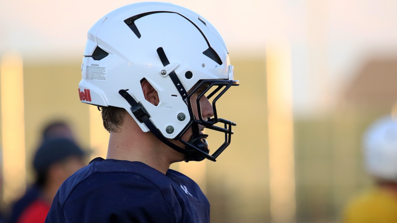Side profile of a football player wearing a white helmet and navy jersey, standing on a practice field.
