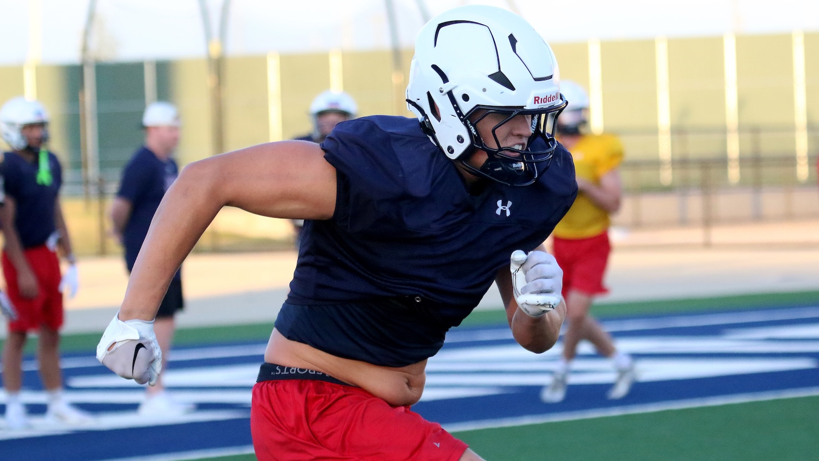 Football player in a navy jersey and white helmet sprints on a blue and green field, teammates blurred in the background.