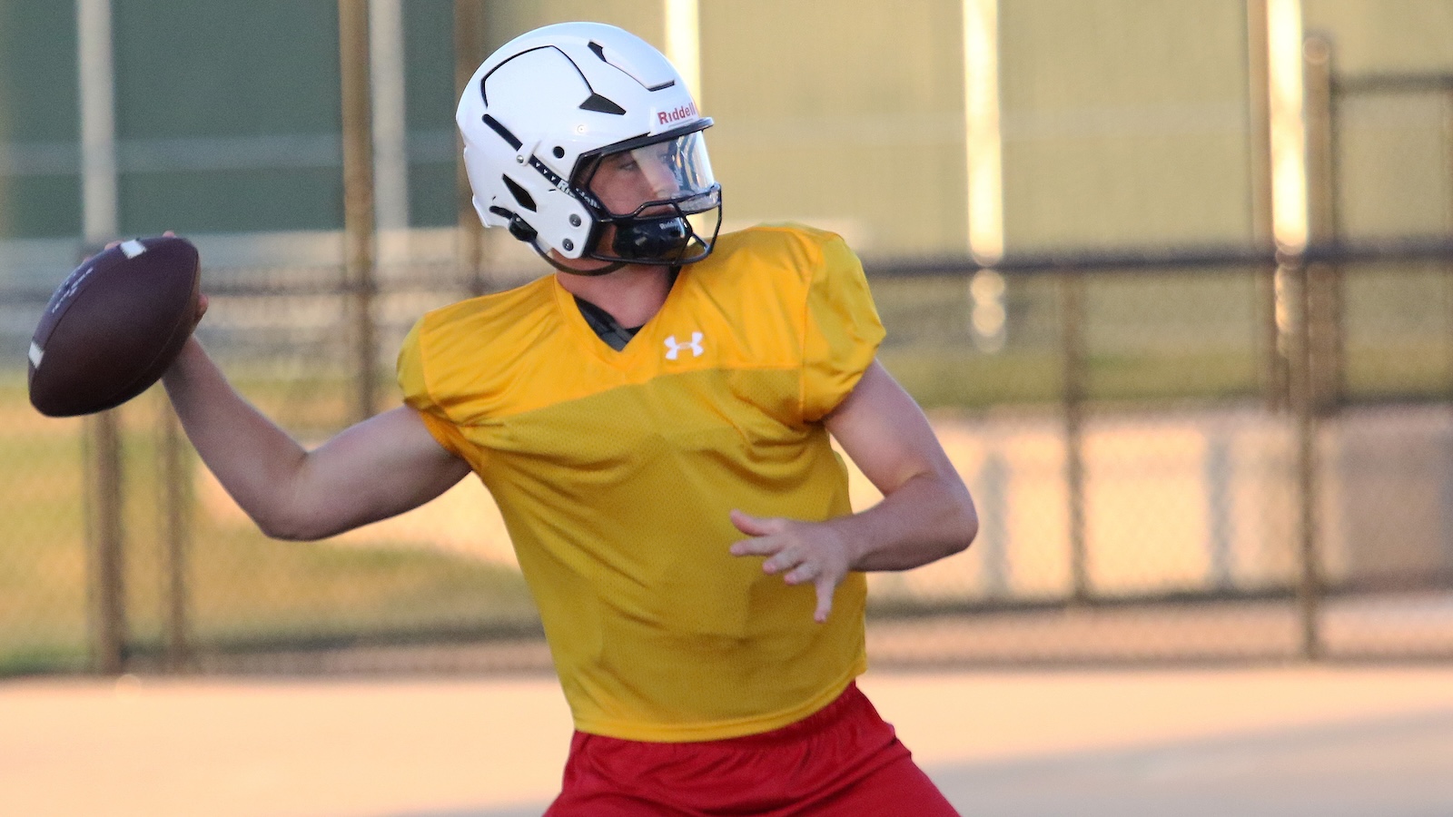 Quarterback in a yellow jersey and white helmet prepares to throw a football on a sunny field where a chain-link fence is visible behind him.