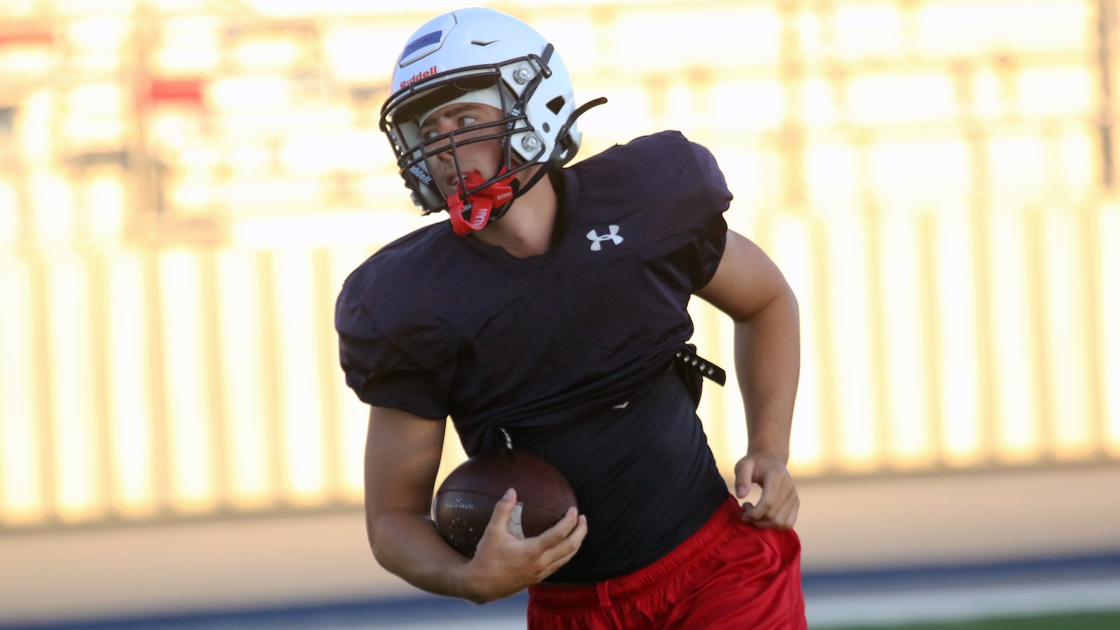 American football player wearing a black jersey and red shorts runs with the ball during practice, wearing a white helmet with a red mouthguard in hand-to-mouth position on the field.