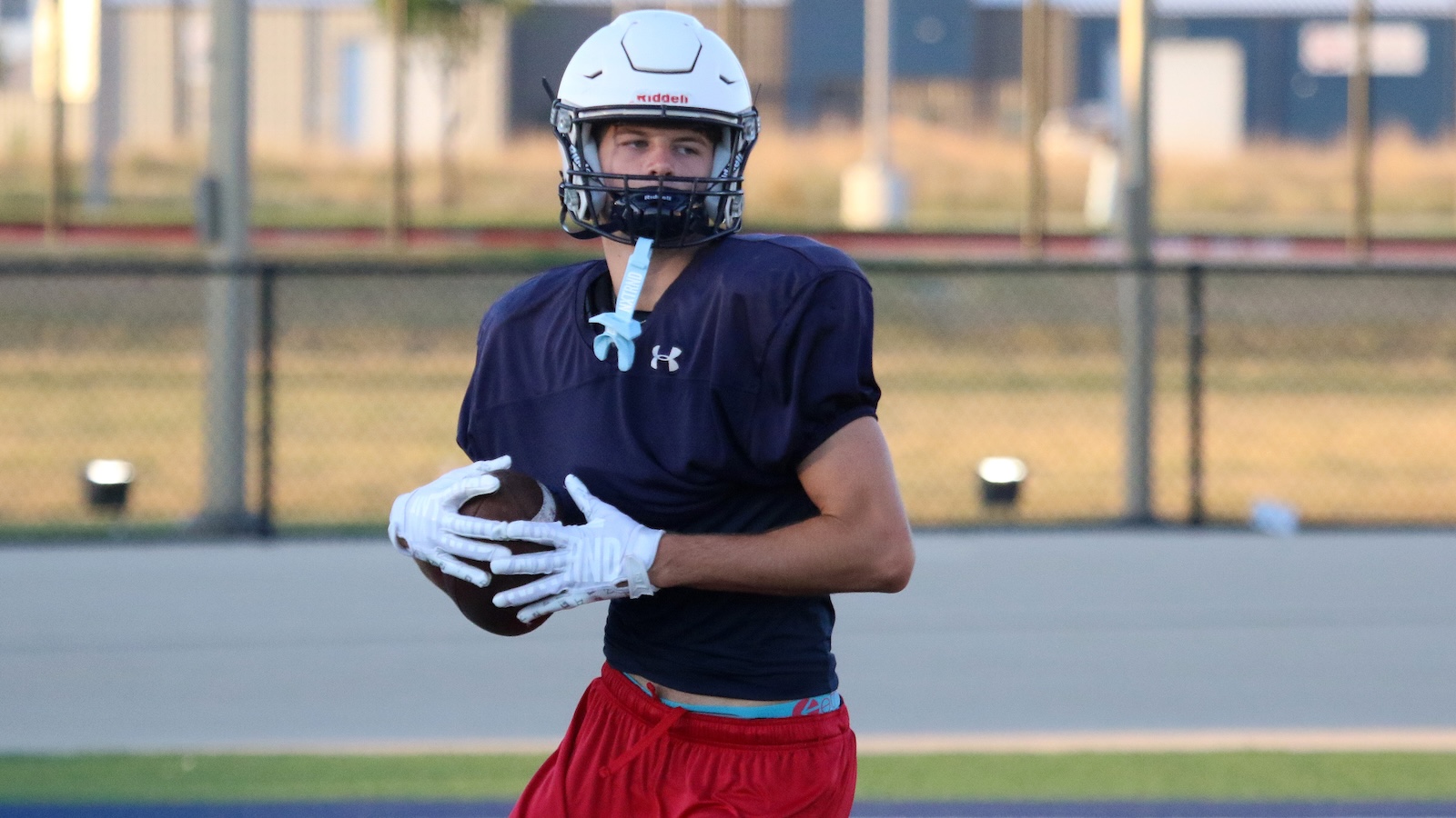 Football player in a navy shirt and red shorts, wearing a white helmet and gloves, holds a football on the field.