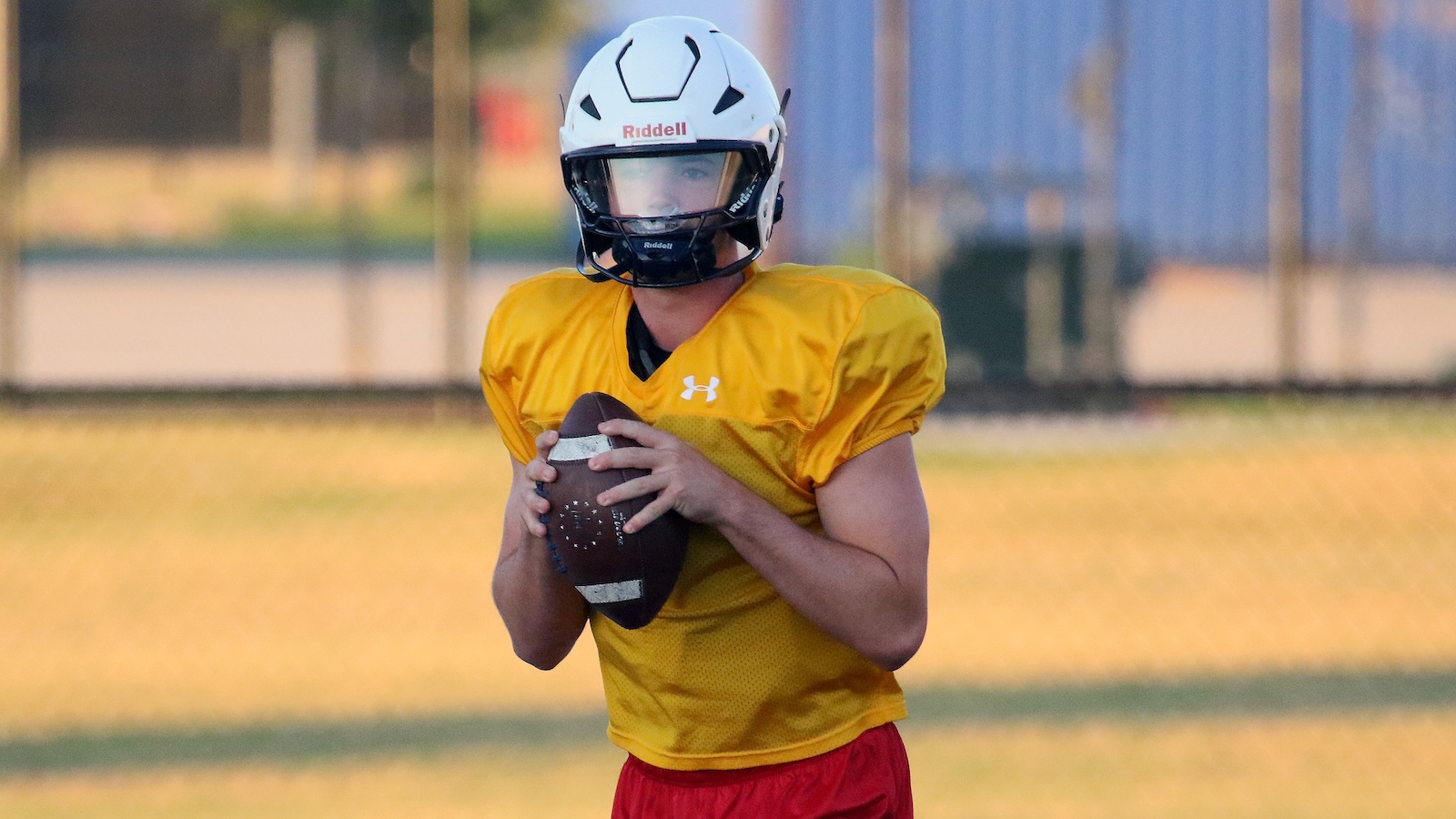 Young football quarterback in a yellow jersey and white helmet grips a football, ready to throw on a field.