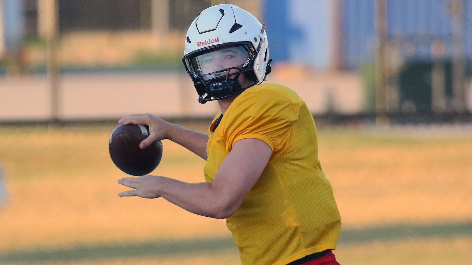 Quarterback in yellow jersey and white helmet prepares to throw a football on a practice field.