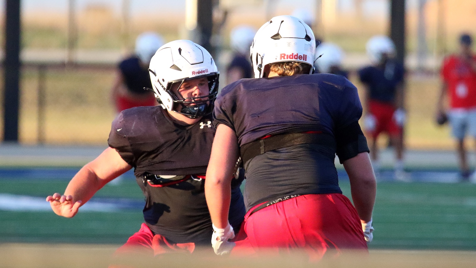 Two football players in black jerseys and white Riddell helmets engage in a drill on a practice field, one facing the other with arms ready.
