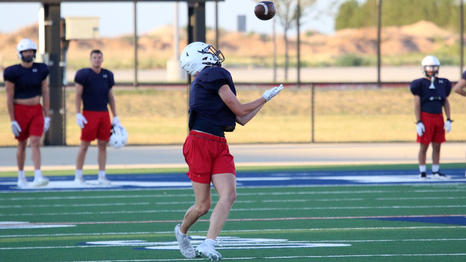 Football practice: player in navy shirt, red shorts and white helmet catches a ball while others watch nearby on a green field.